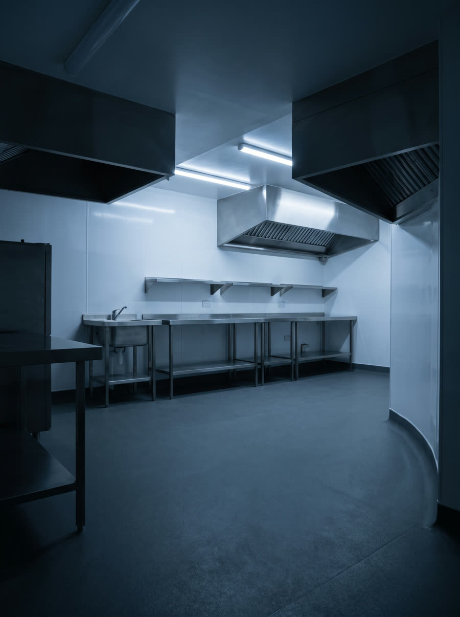 An empty commercial kitchen with stainless benching against clad walls and a coved floor junction.